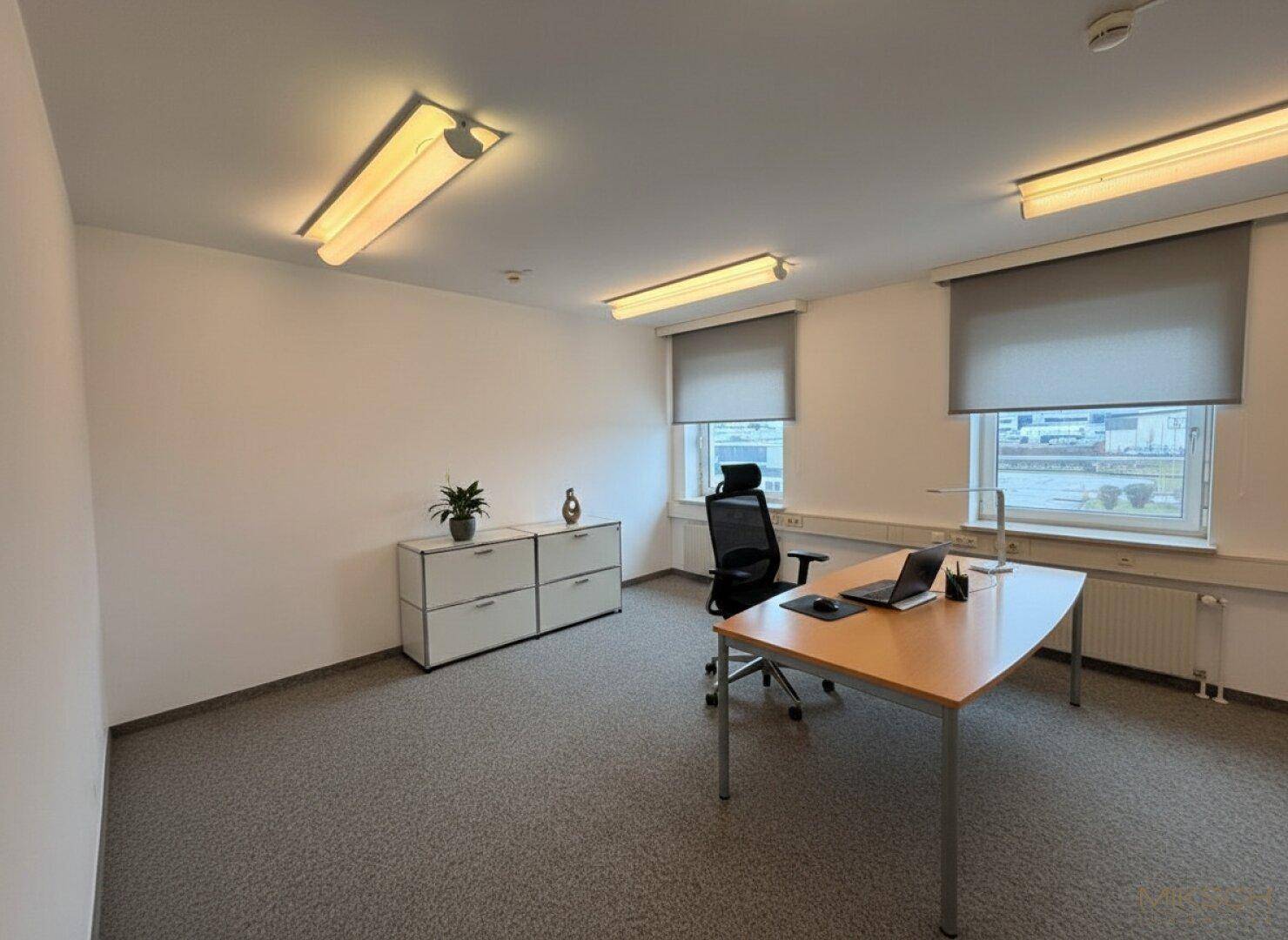 Einzelbüro mit Teppichboden, modernem Schreibtisch und zwei Fenstern mit Rollos.