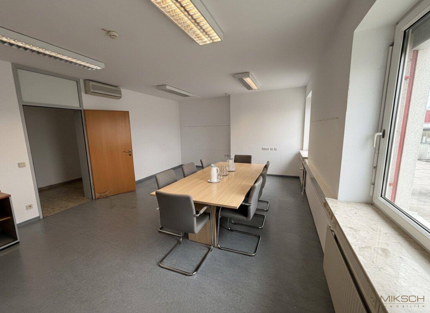 Besprechungsraum mit großem Holztisch, grauen Stühlen und Fenstern mit Blick nach draußen.