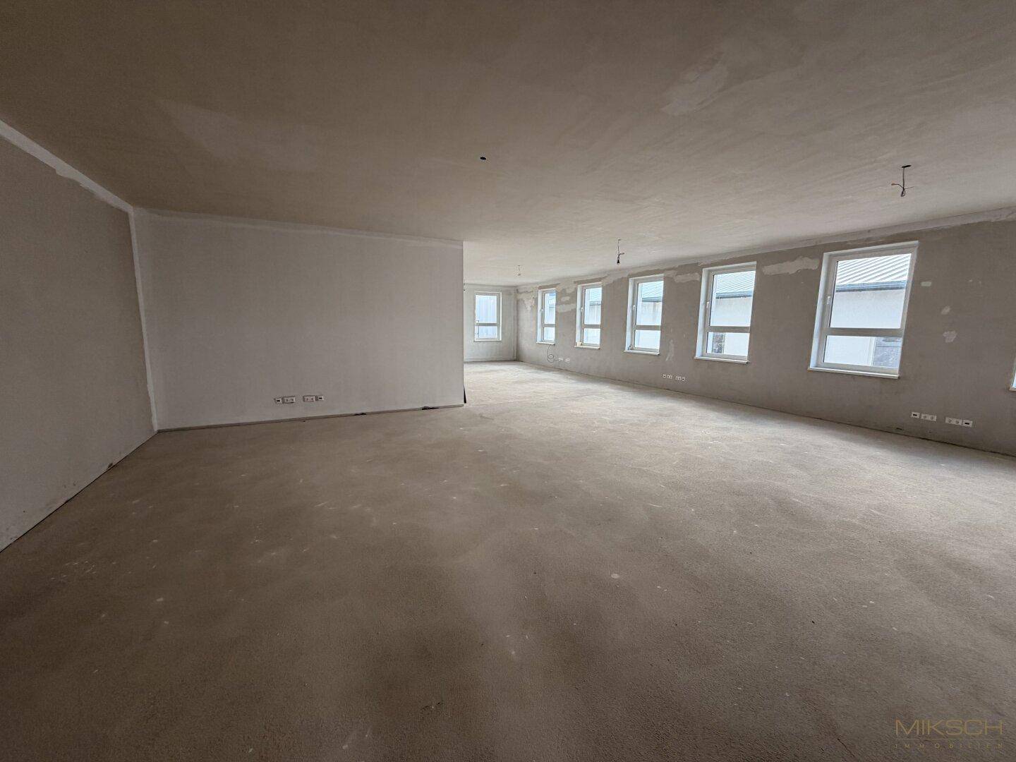 Großer, leerer Raum im Rohbauzustand mit vielen Fenstern und Betonboden.