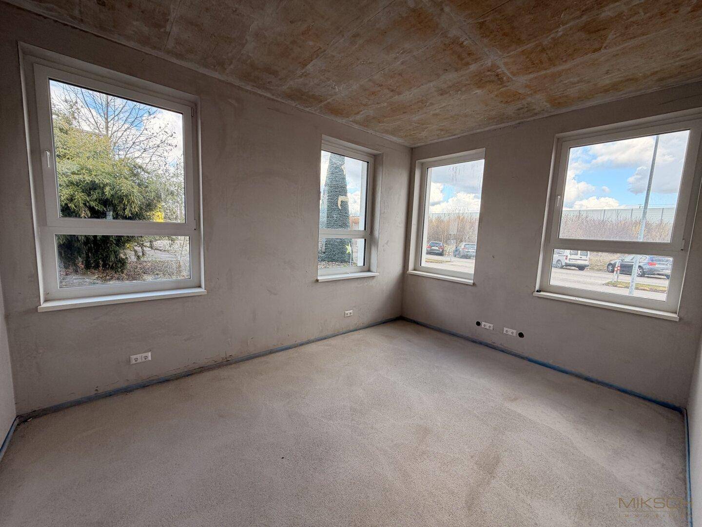 Eckraum im Rohbau mit mehreren Fenstern, Betonboden und Blick nach draußen.