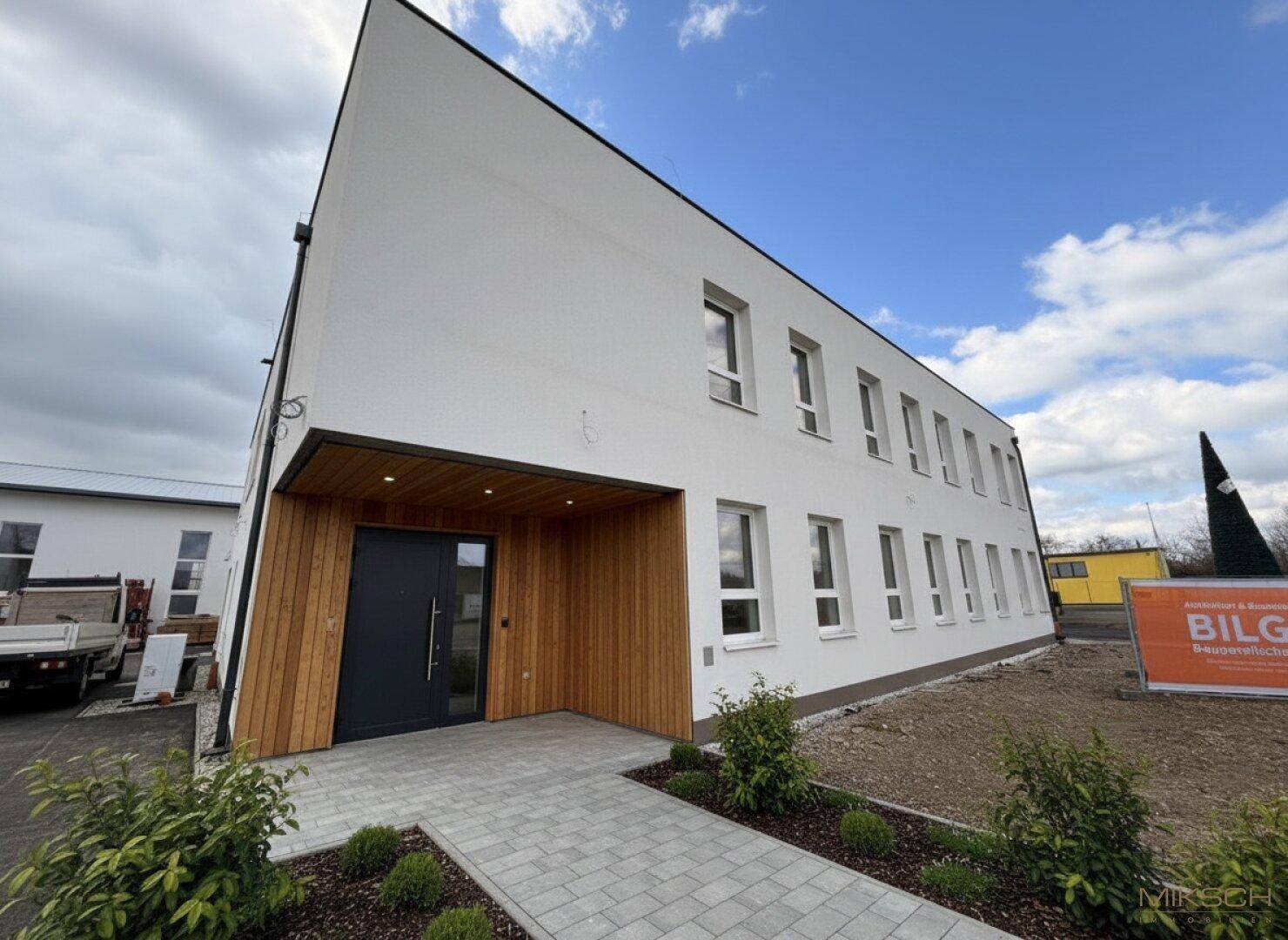 Moderne Geschäftsfassade mit weißem Putz, Holzakzenten am Eingang und gepflastertem Zugangsbereich.