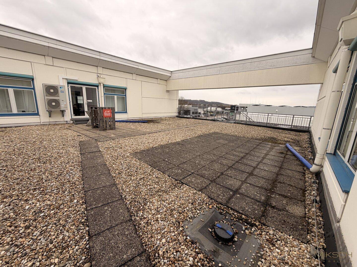 Dachterrasse mit Kies und Betonplatten, umgeben von einer Brüstung und Blick auf die Umgebung.