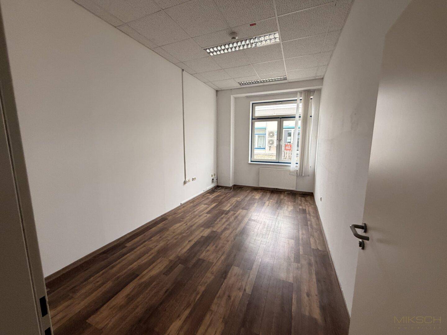 Helles Einzelbüro mit dunklem Laminatboden, weißen Wänden und einem Fenster mit Blick nach draußen.