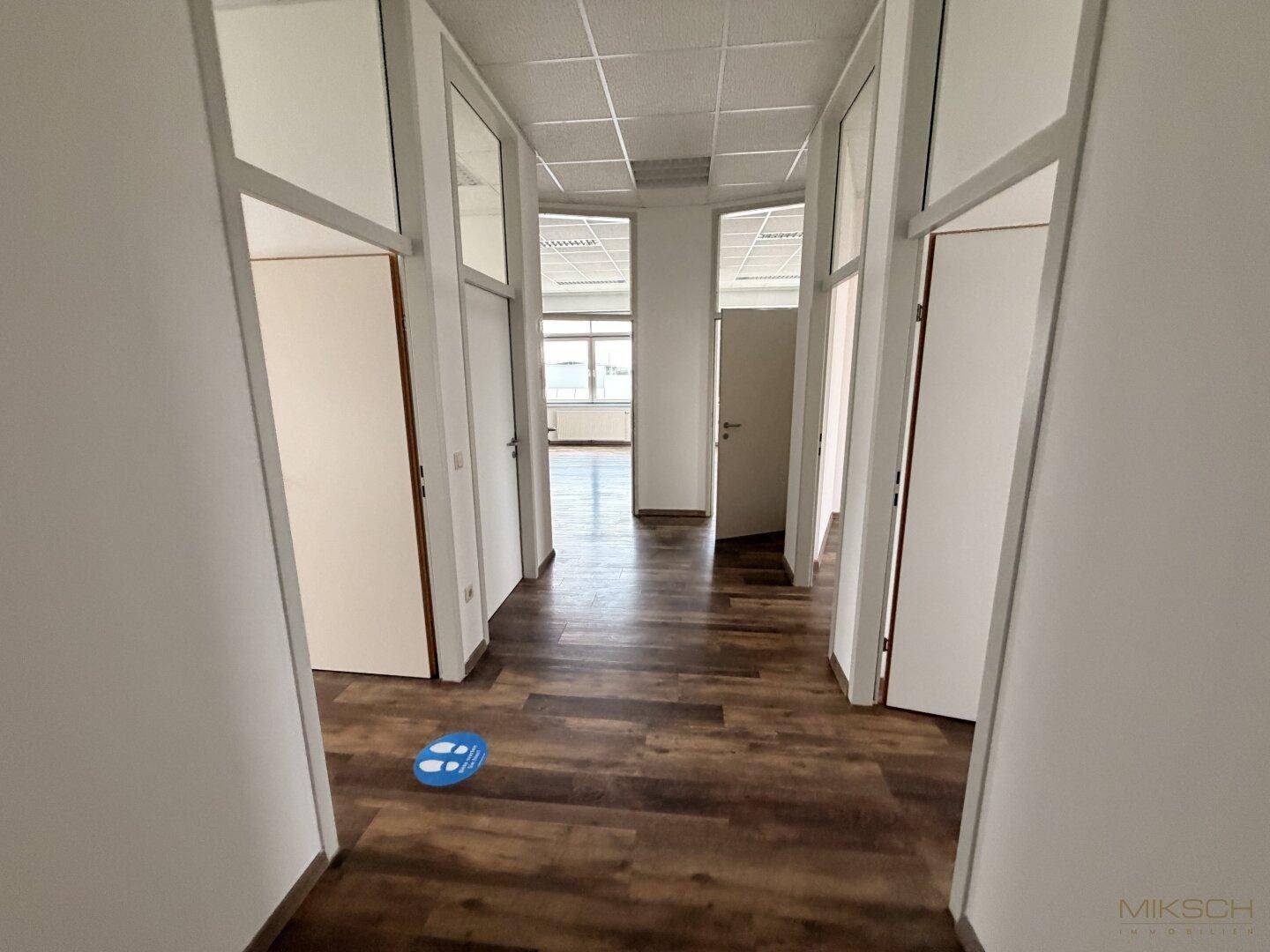 Flur mit dunklem Laminatboden und weißen Wänden, der zu Büros mit Glastüren und einem offenen Bereich führt.
