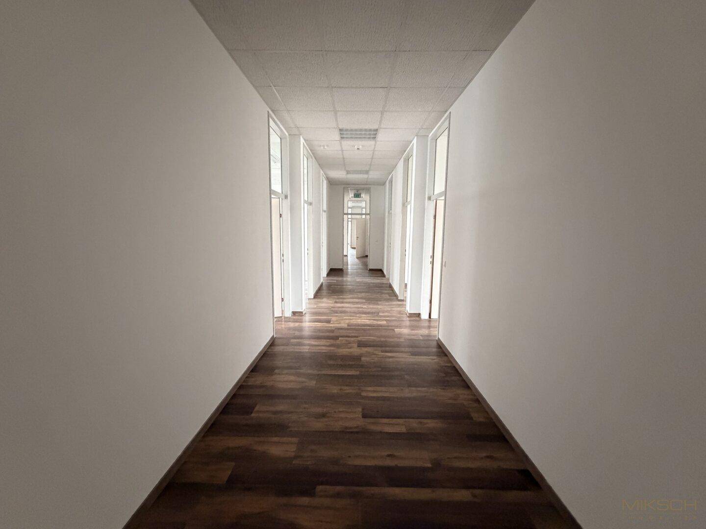 Langer Flur mit dunklem Laminatboden und weißen Wänden, der zu weiteren Büroräumen führt.