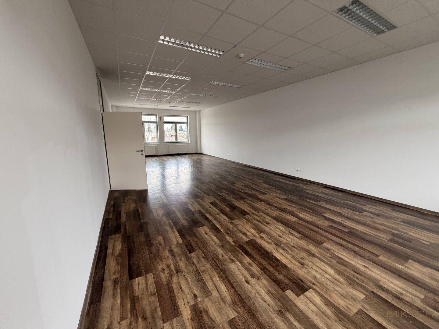 Großes, leeres Büro mit dunklem Laminatboden, weißen Wänden und Fenstern, die Tageslicht spenden.