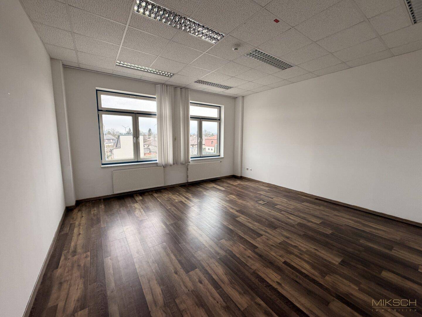 Heller Büroraum mit dunklem Laminatboden, weißen Wänden und zwei Fenstern mit Vorhängen.