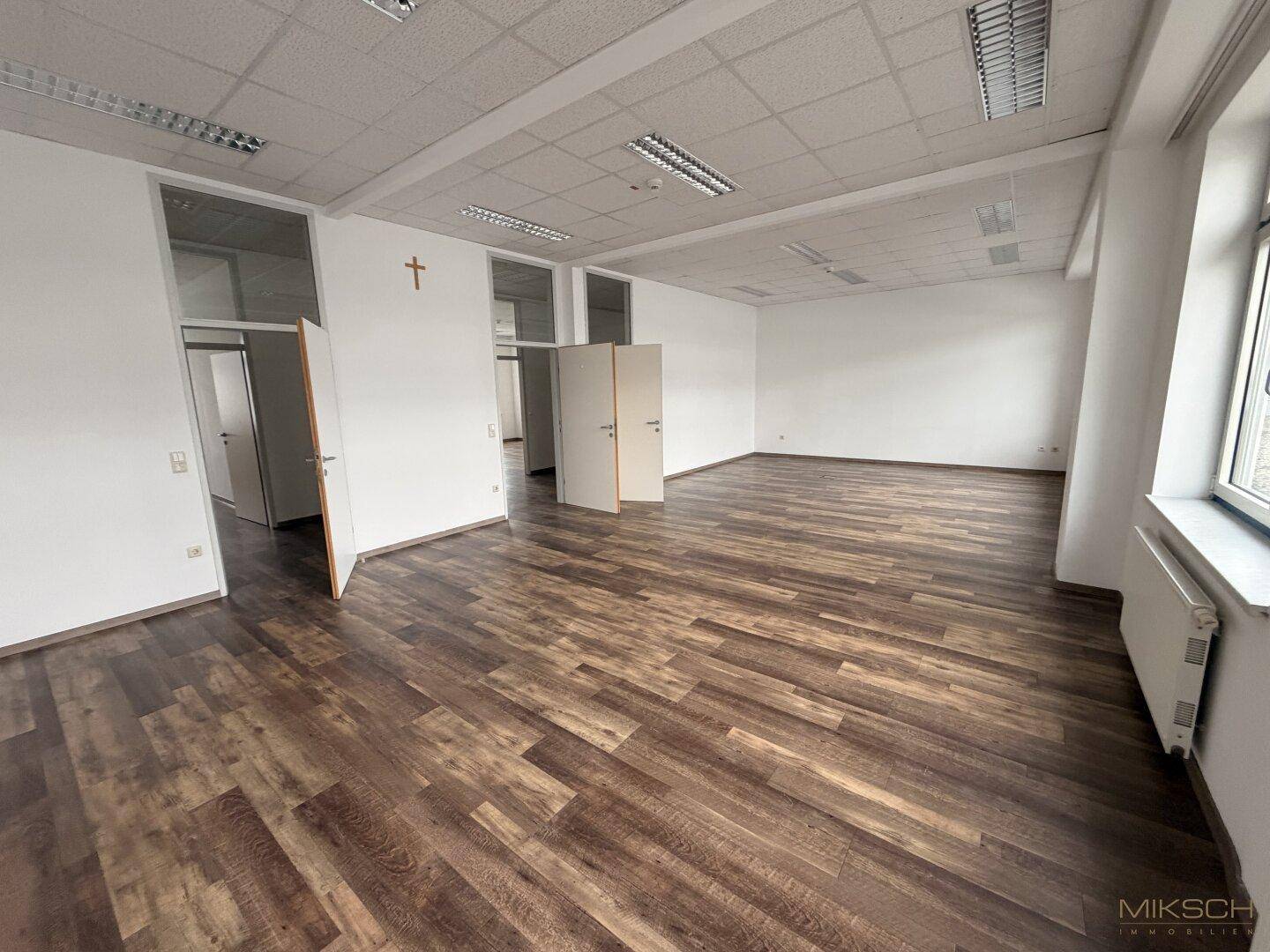 Offener Bürobereich mit dunklem Laminatboden, weißen Wänden und mehreren Türen zu angrenzenden Räumen.