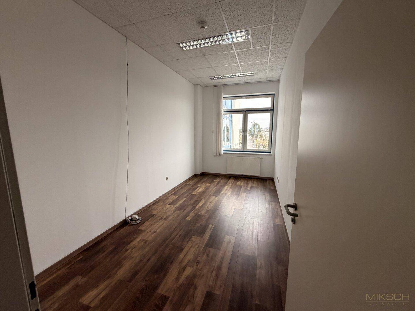 Kleineres Büro mit dunklem Laminatboden, weißen Wänden und einem Fenster, das für natürliches Licht sorgt.