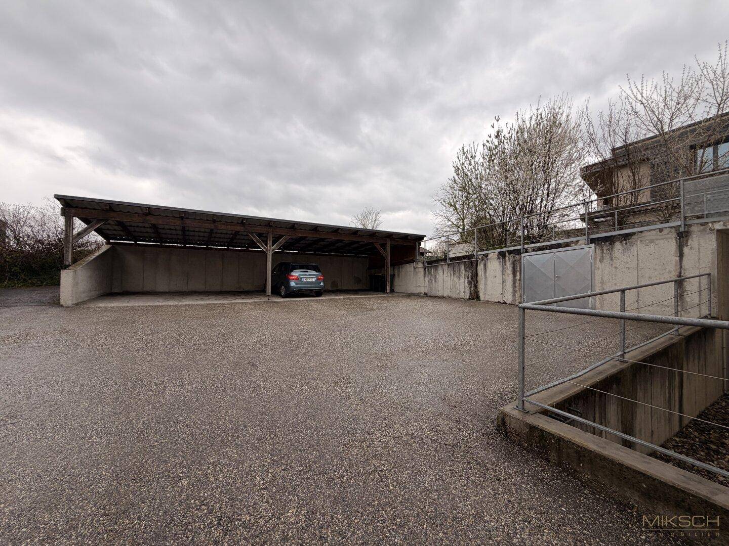 Überdachter Stellplatz und offener Parkbereich mit einem Auto unter einem Carport an einem bewölkten Tag.