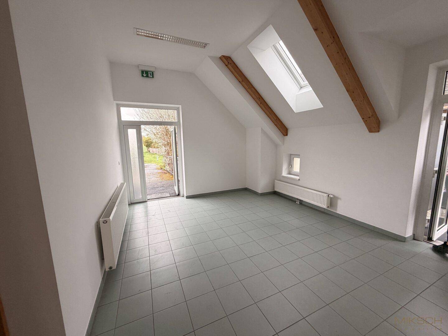 Heller Eingangsbereich mit Fliesenboden, Dachschräge und Zugang zu einem Außenbereich durch eine Glastür.