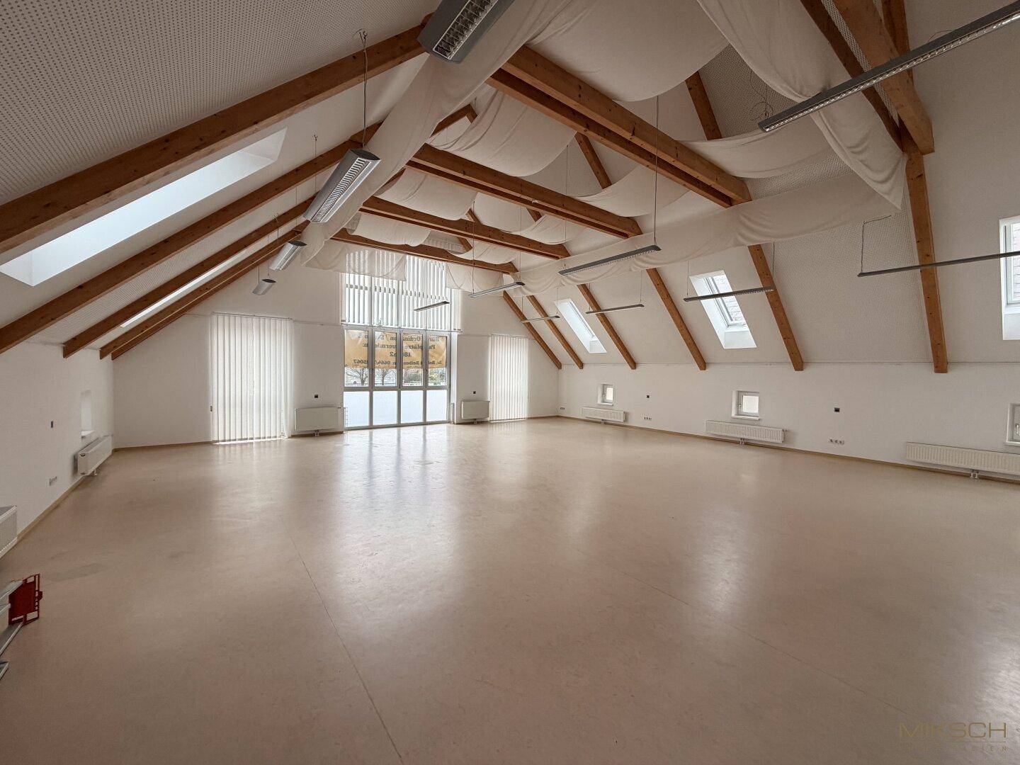 Geräumiger, heller Dachgeschossraum mit sichtbaren Holzbalken, vielen Dachfenstern und hellem Bodenbelag.