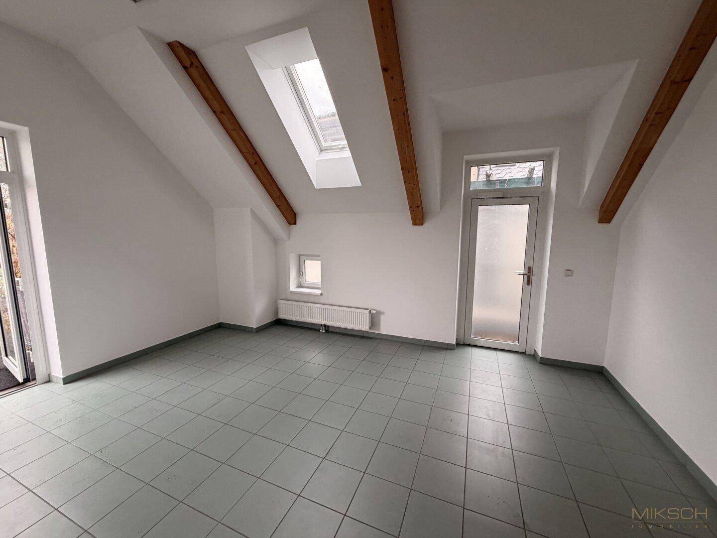 Heller Raum mit Fliesenboden, Dachschräge und zwei Türen, die nach draußen oder in andere Bereiche führen.