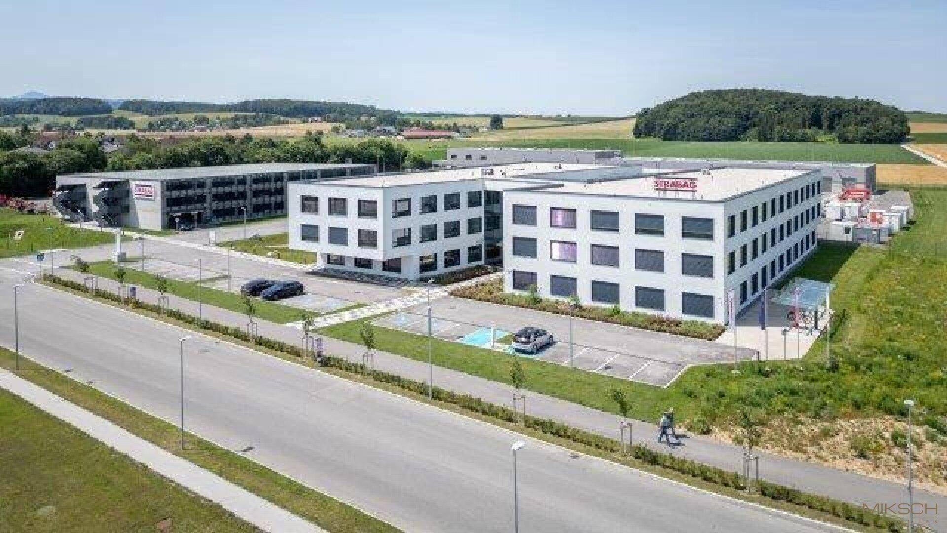 Gesamtansicht eines modernen Bürokomplexes mit Parkplätzen, umgeben von grüner Landschaft und einer Straße.