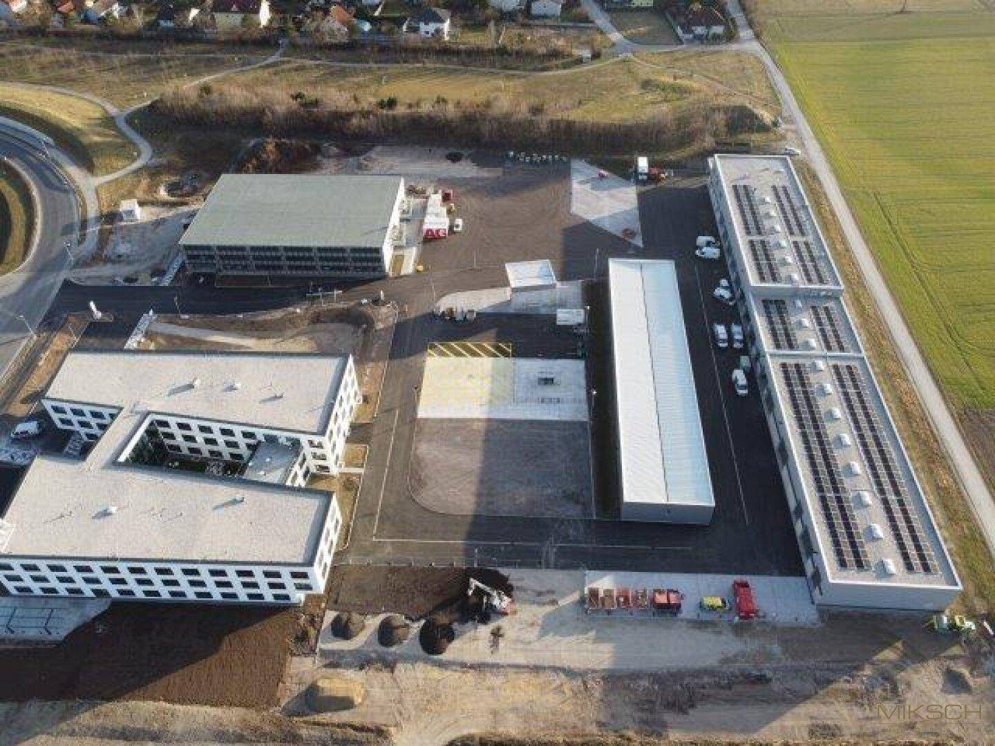 Luftaufnahme eines weitläufigen Gewerbegebiets mit mehreren Gebäuden, Parkflächen und Solaranlagen auf Dächern.