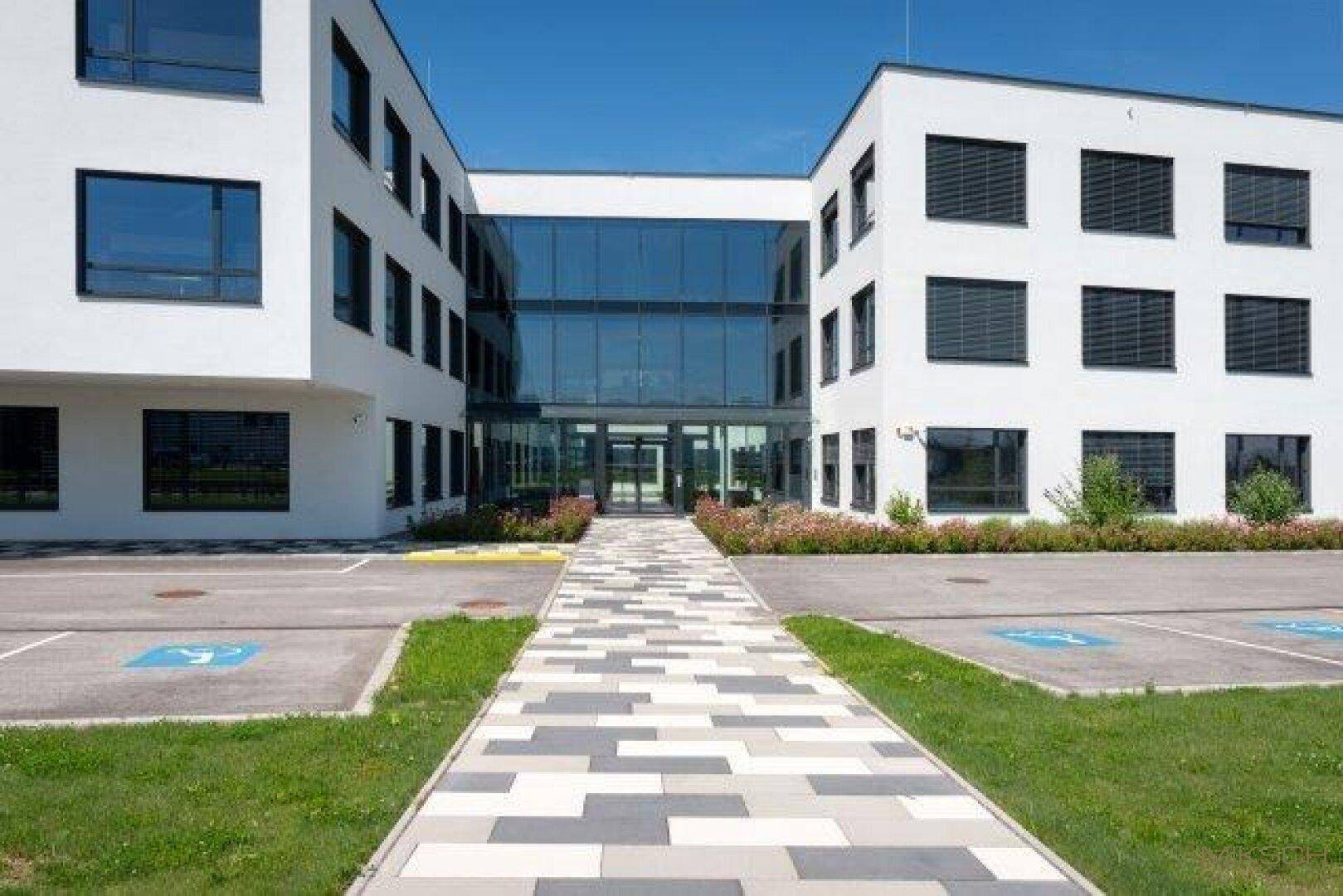Modernes Bürogebäude mit weißer Fassade, großen Fenstern und einem gepflasterten Eingangsbereich.