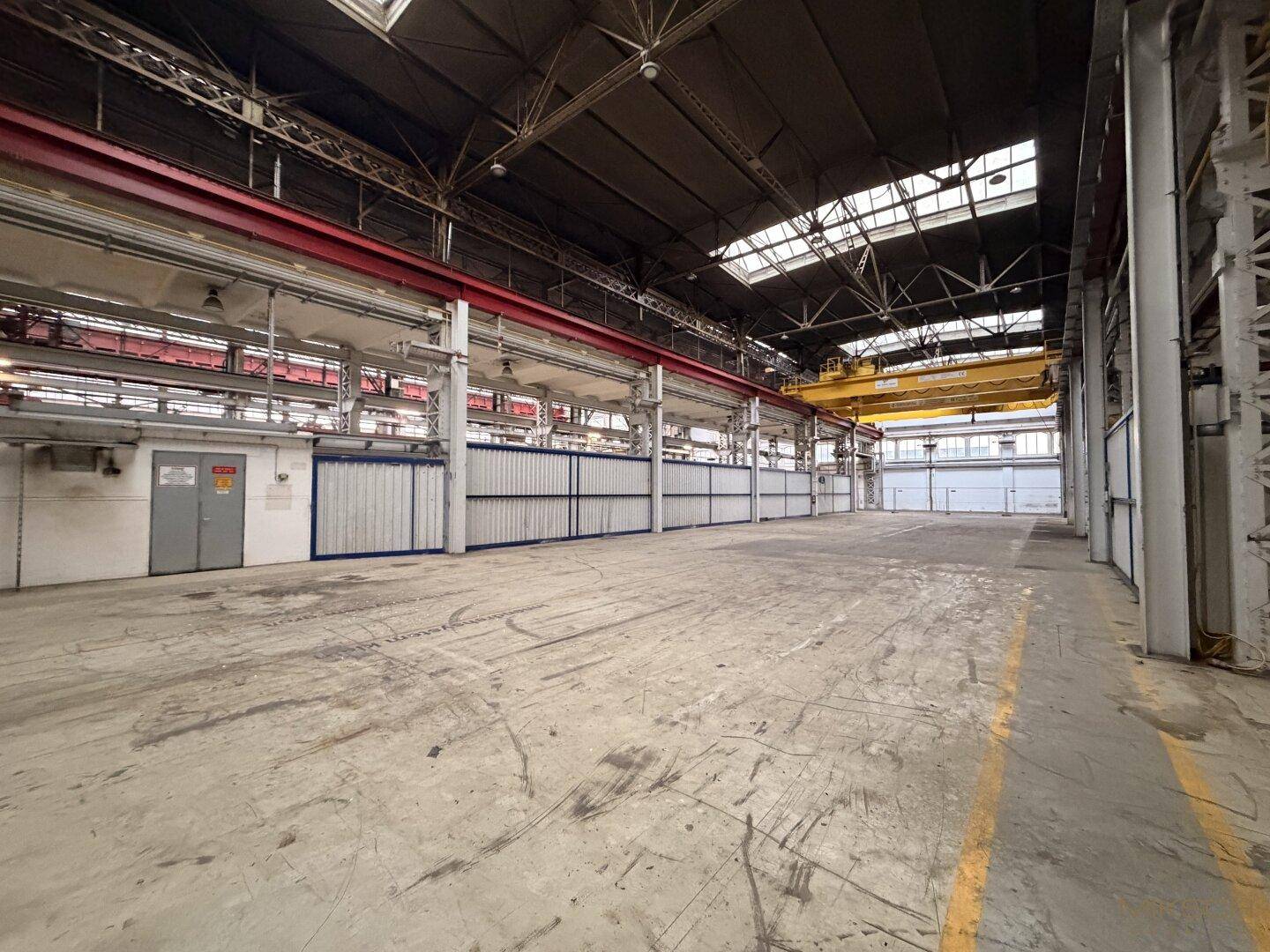 Weitläufige Industriehalle mit Betonboden, hohen Decken und einem gelben Brückenkran, bereit für industrielle Nutzung.