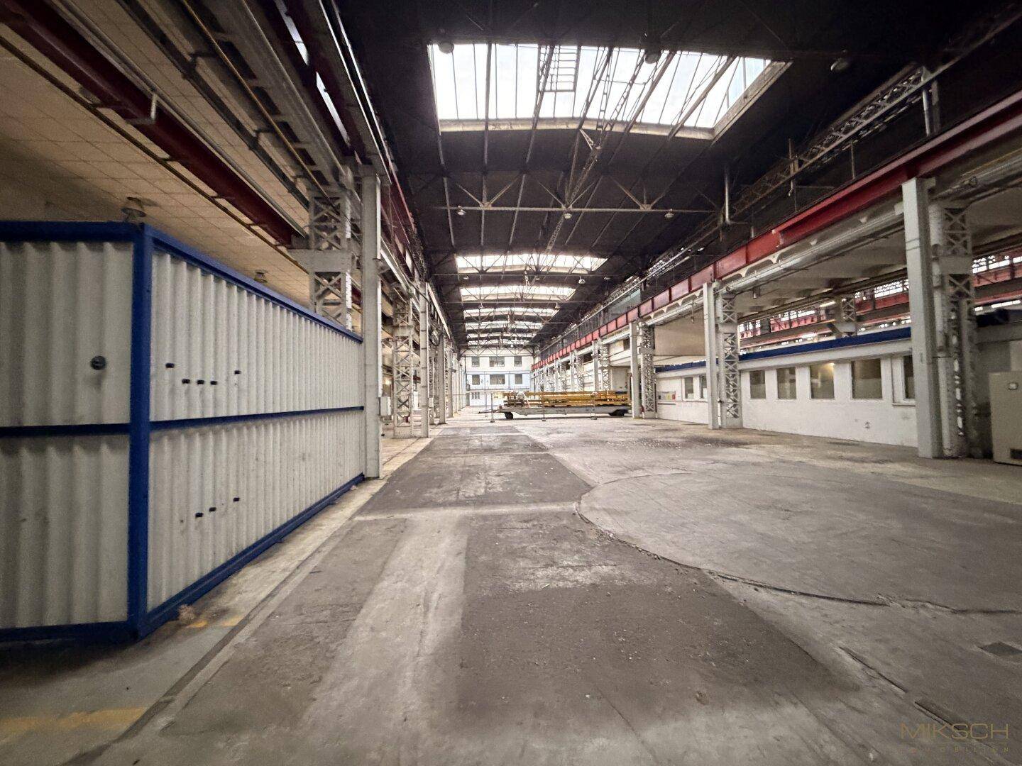 Langer Gang in einer Industriehalle mit Betonboden, hohen Decken und einem blauen Container an der Seite.