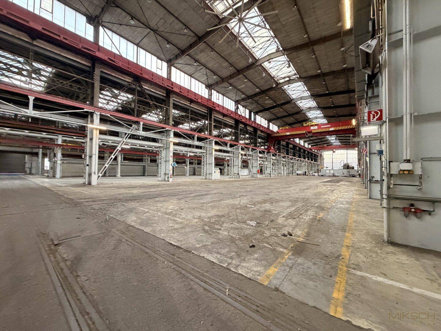 Weitläufige Industriehalle mit hohen Decken, Betonboden und sichtbaren roten Kranbahnen, bietet viel Platz.