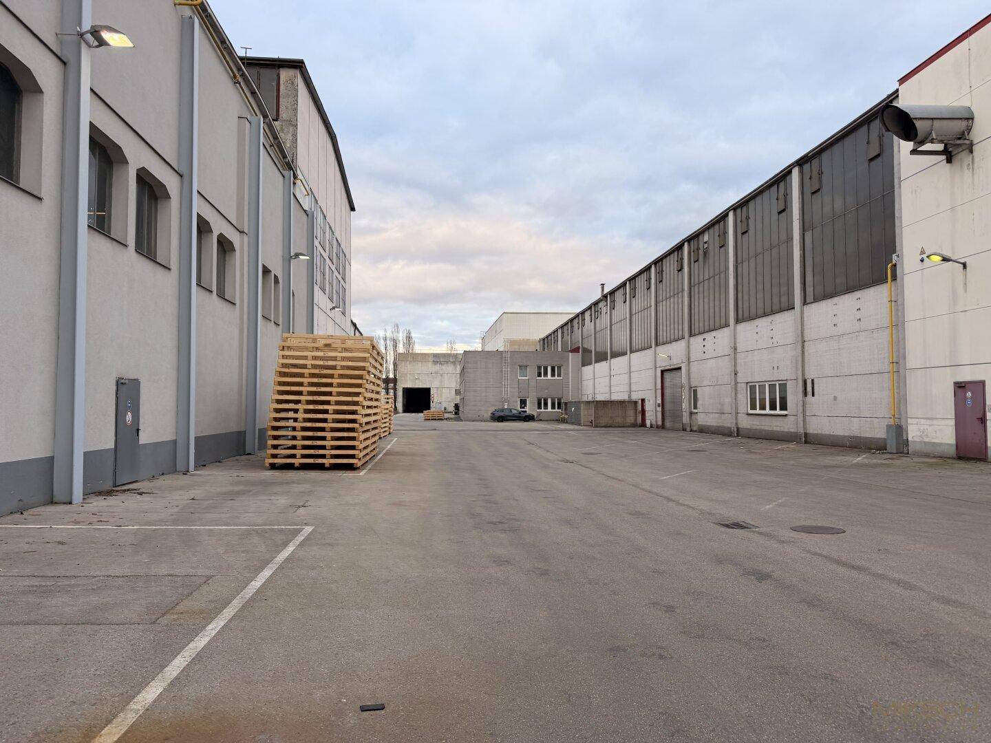 Außenbereich zwischen zwei Industriegebäuden mit asphaltiertem Hof und gestapelten Holzpaletten, ideal für Logistik.