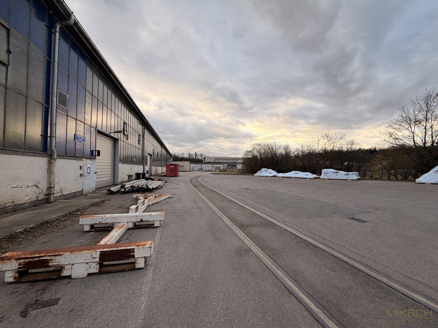 Außenbereich mit Bahngleisen und einer langen Fassade eines Industriegebäudes unter einem bewölkten Himmel.