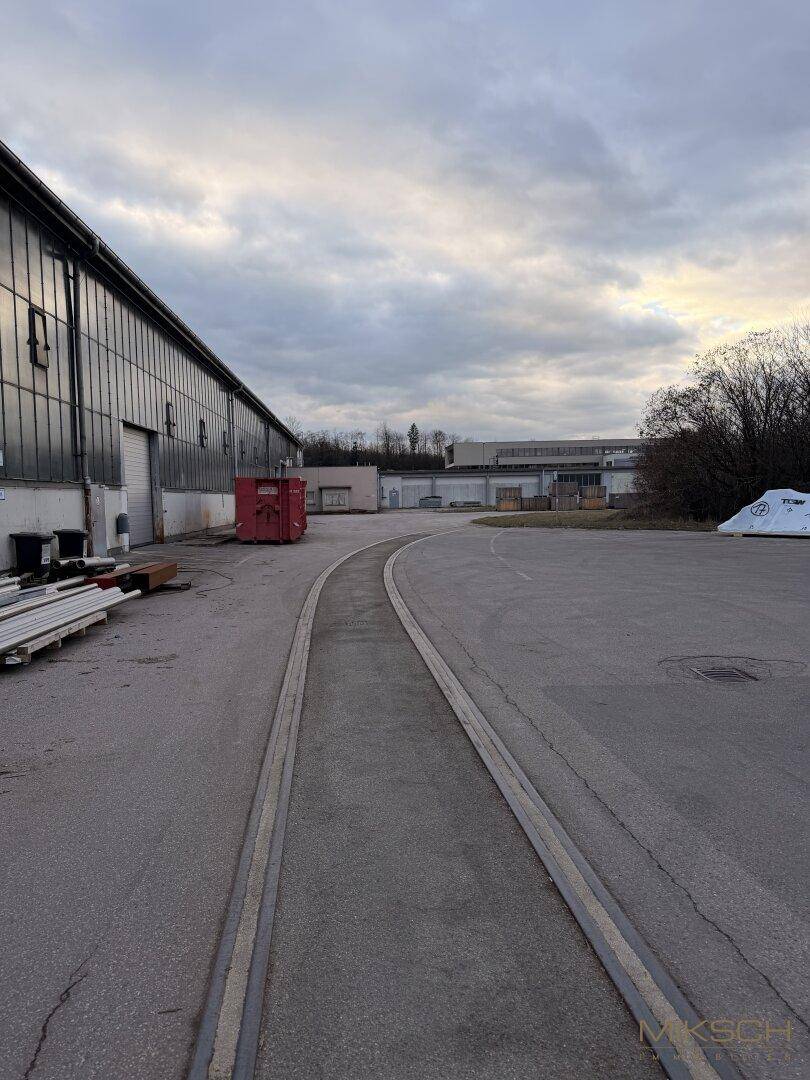 Außenansicht mit alten Bahngleisen auf asphaltiertem Gelände neben einem Industriegebäude unter bewölktem Himmel.