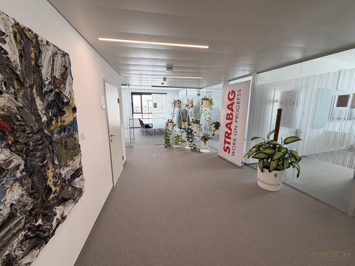 Heller Bürogang mit Kunst an der Wand, Pflanzen und einem "STRABAG" Banner, der den Arbeitsbereich kennzeichnet.