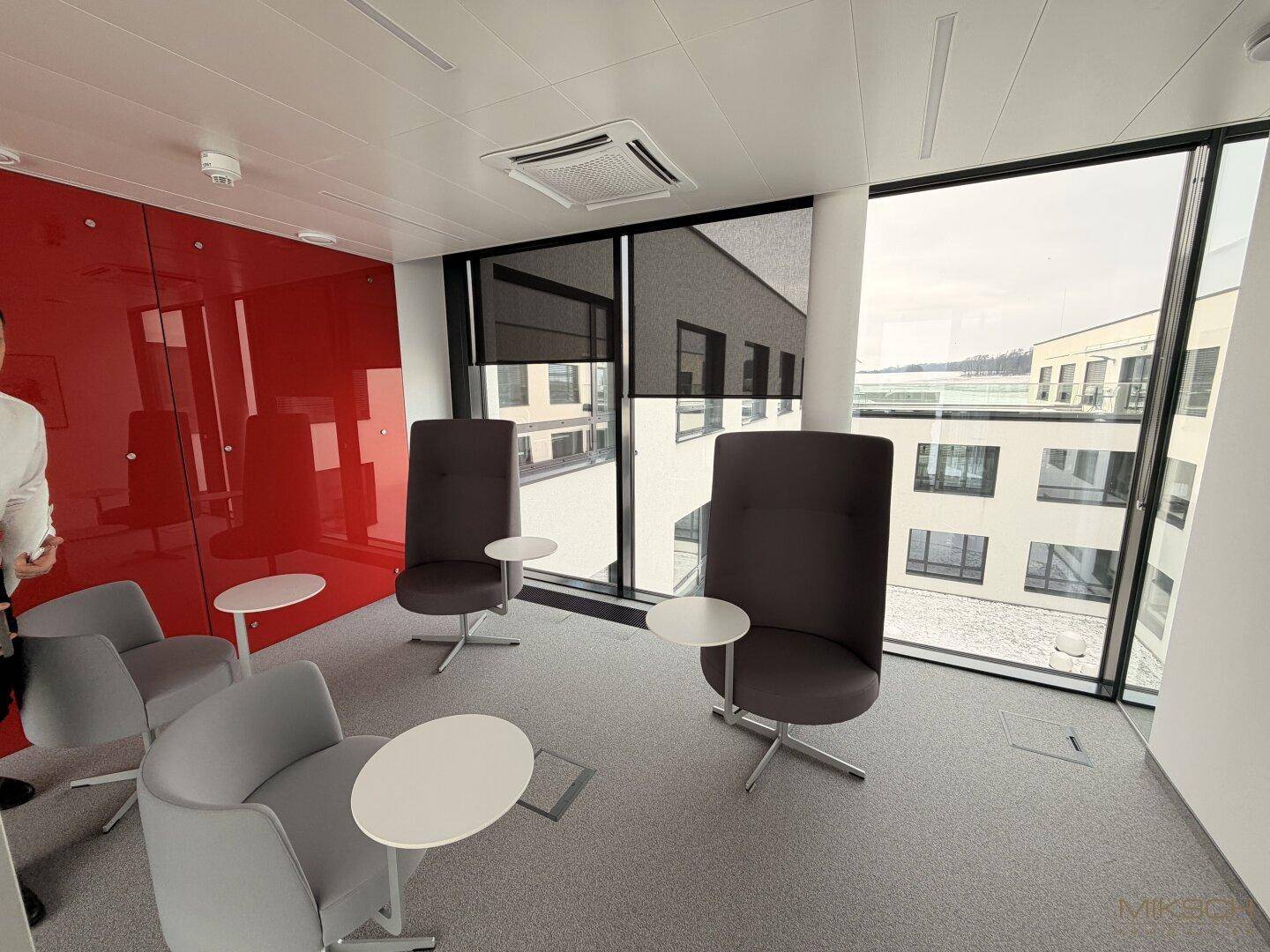 Moderne Lounge-Ecke mit roten Akzentwänden, bequemen Sesseln und großen Fenstern mit Blick nach draußen.