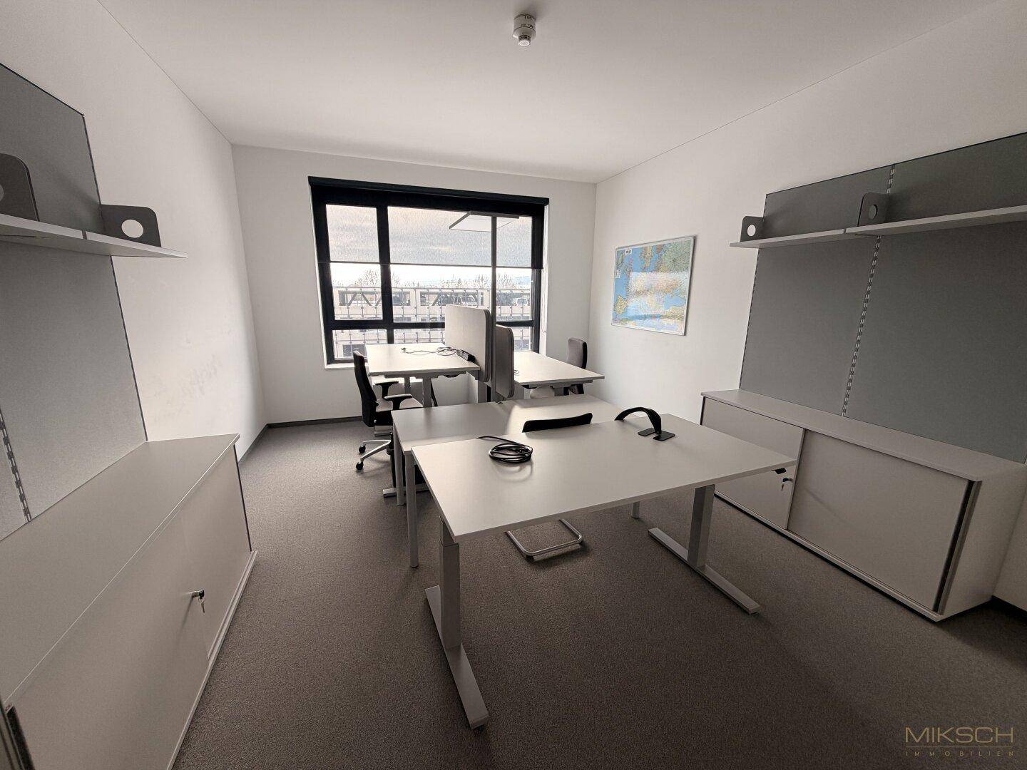 Helles Einzelbüro mit zwei Schreibtischen, Bürostühlen und einem großen Fenster mit Blick auf die Umgebung.