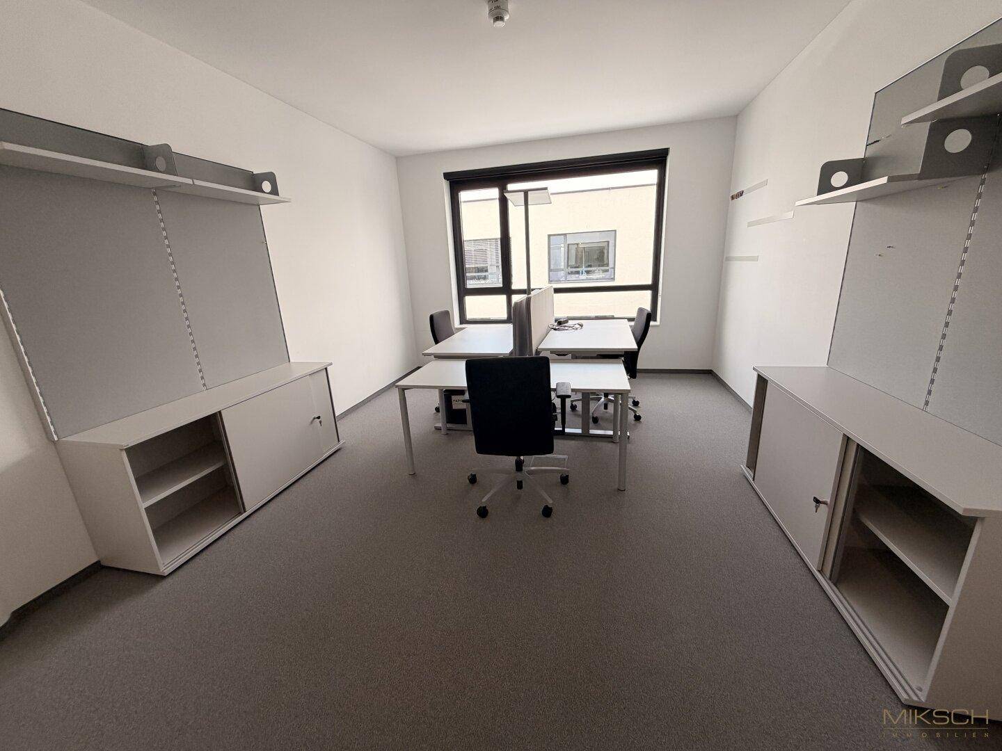 Geräumiges Büro mit zwei Arbeitsplätzen, weißen Schränken und einem Fenster, das Tageslicht hereinlässt.