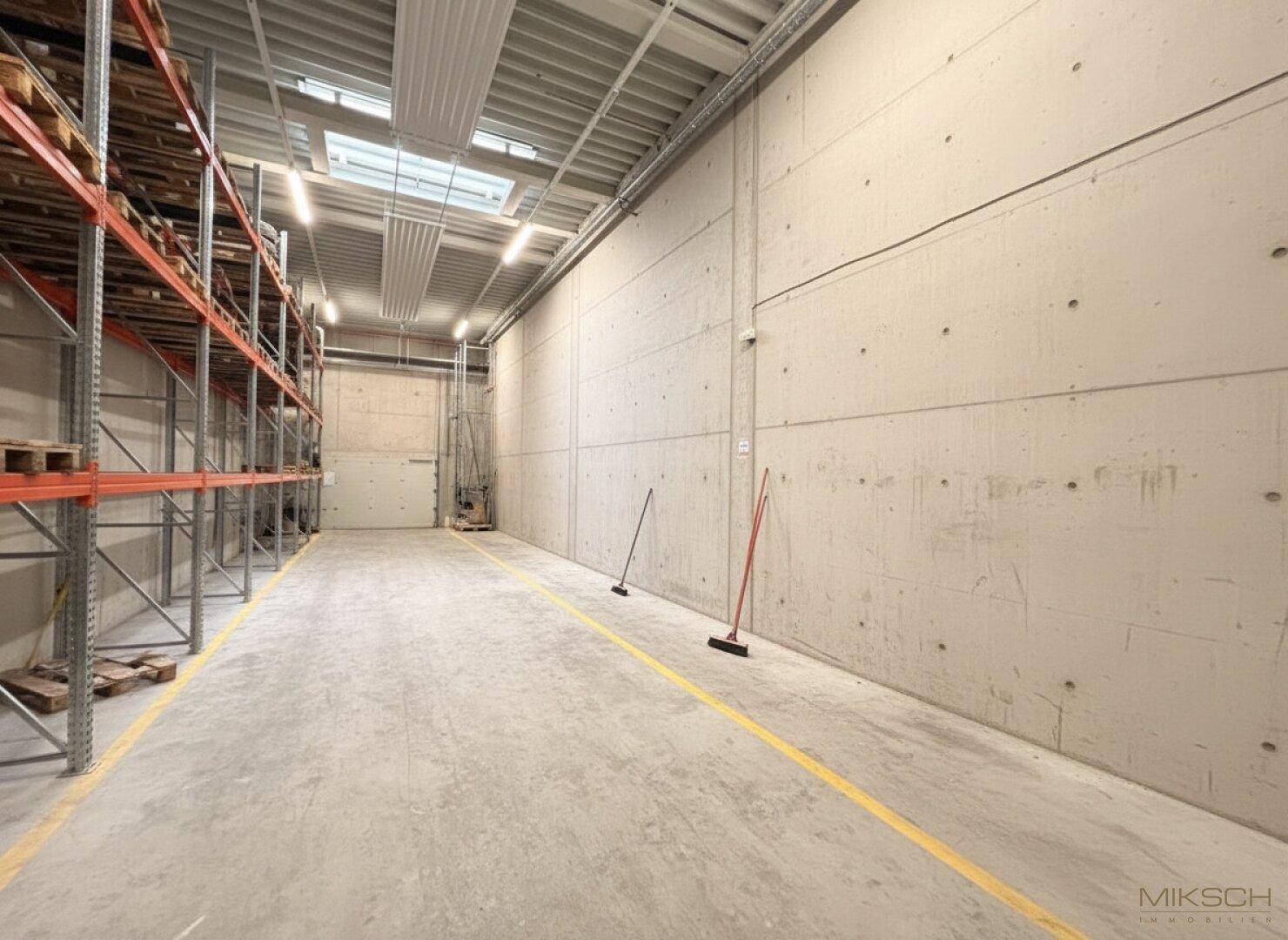 Geräumige Lagerhalle mit hohen Decken, Betonwänden, Regalsystemen und einem Betonboden mit Markierungen.