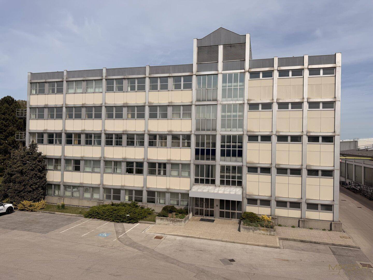 Luftaufnahme des Geschäftsgebäudes mit seiner Fassade und dem umliegenden Parkplatz, eingebettet in die Umgebung.