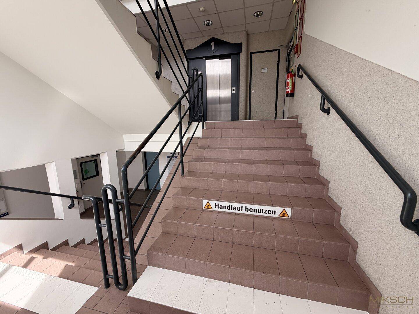 Treppenhaus mit braunen Fliesen, schwarzem Geländer und einem Aufzug, der die Etagen verbindet.