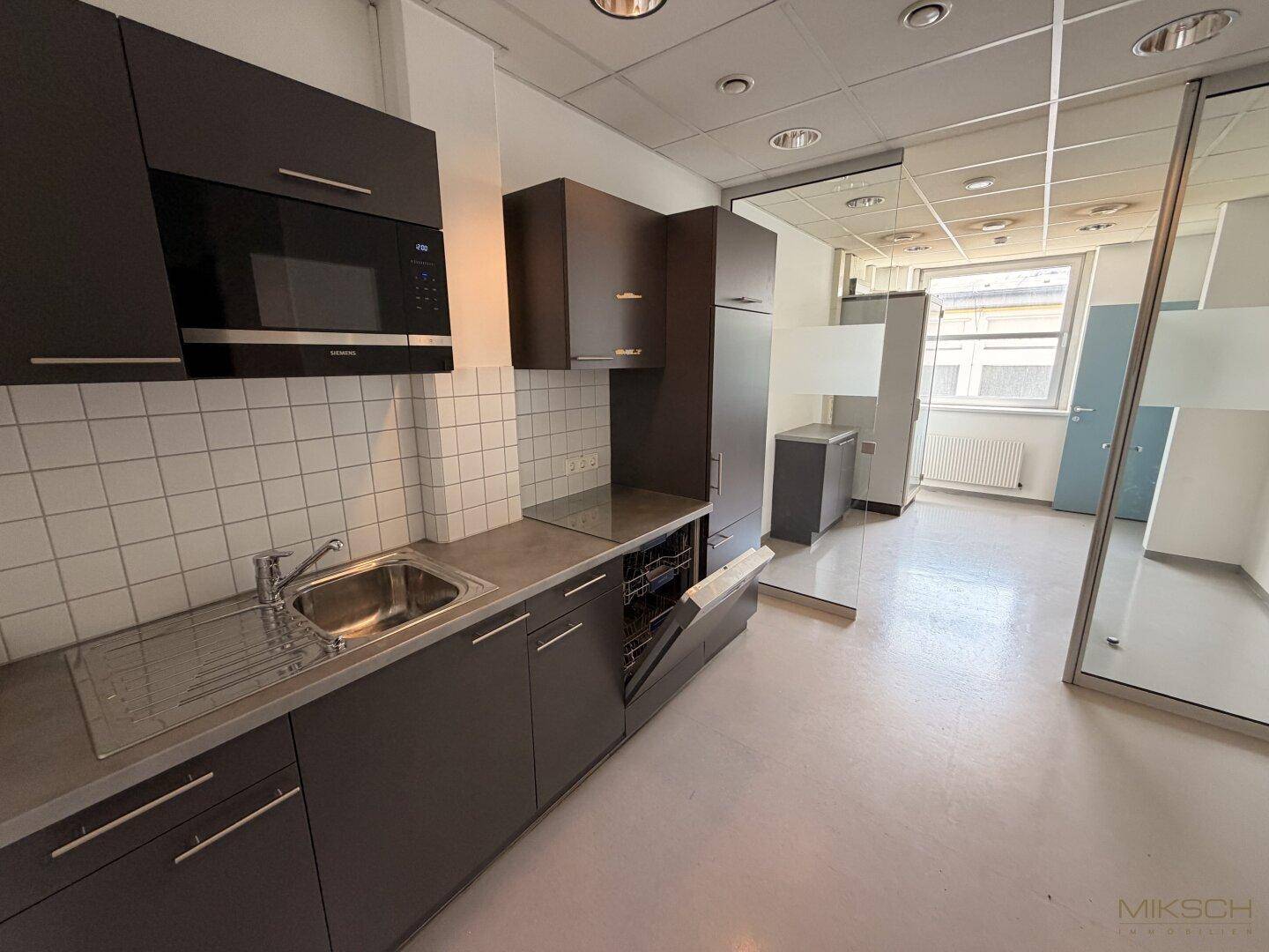 Moderne Küchenzeile mit dunklen Schränken, Spülmaschine und Mikrowelle, ideal für eine Büroumgebung.