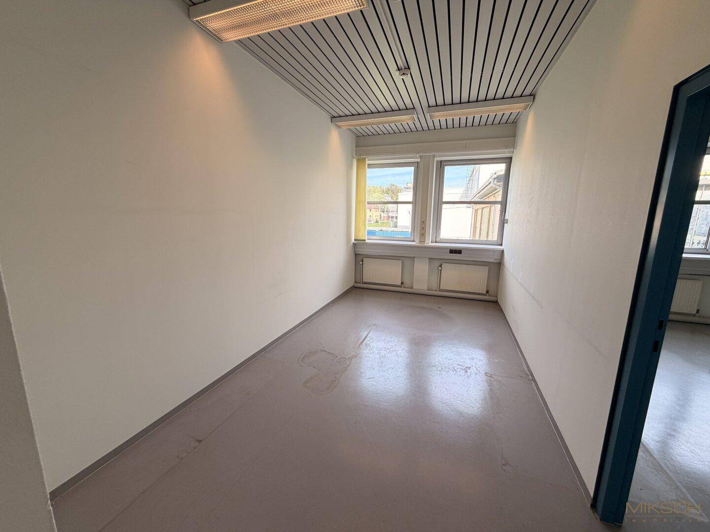 Leerer Büroraum mit hellgrauem Boden und zwei Fenstern, bereit zur individuellen Gestaltung.