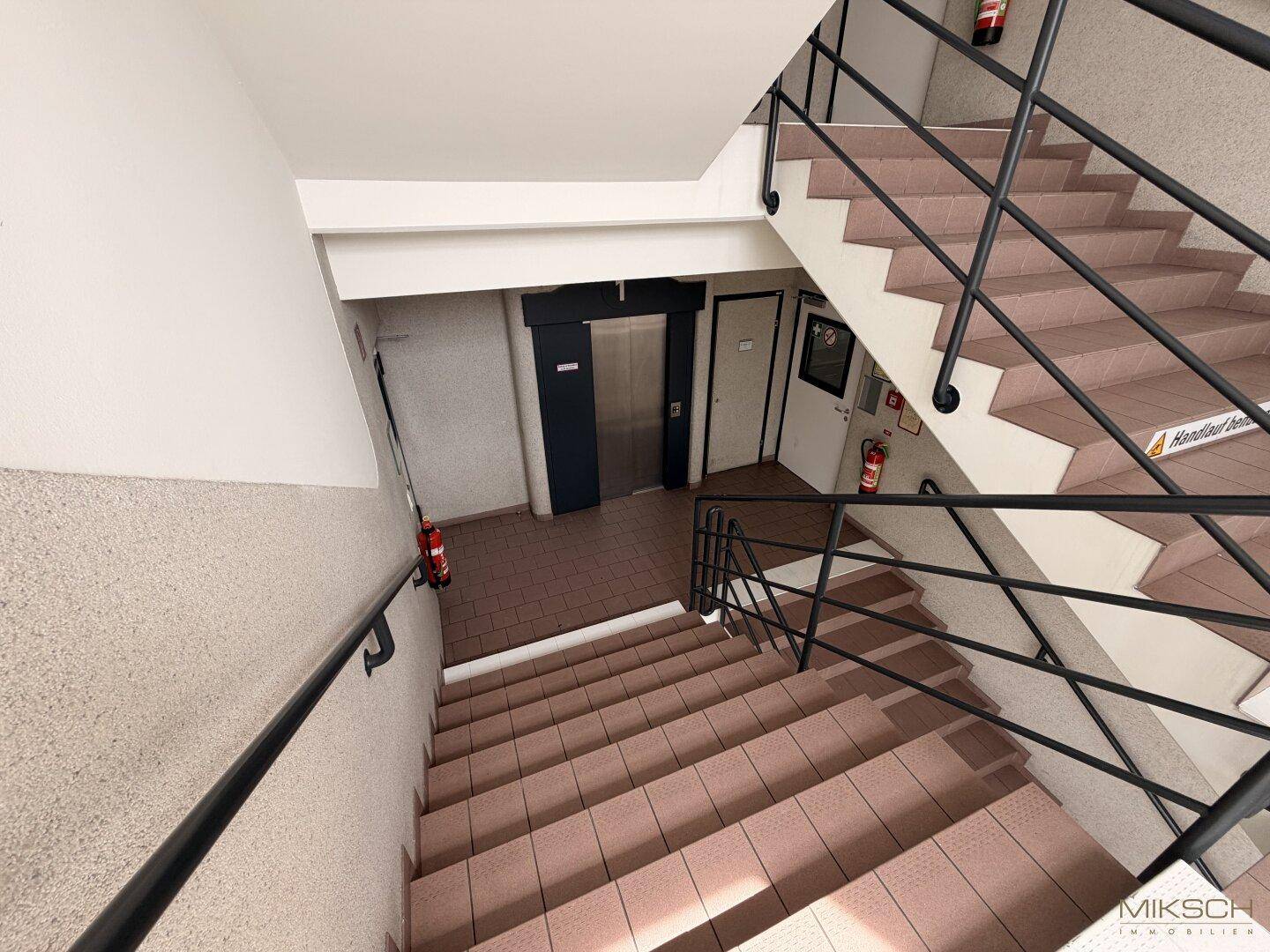 Geräumiges Treppenhaus mit Fliesenboden und schwarzem Metallgeländer, das auch einen Aufzugsbereich zeigt.