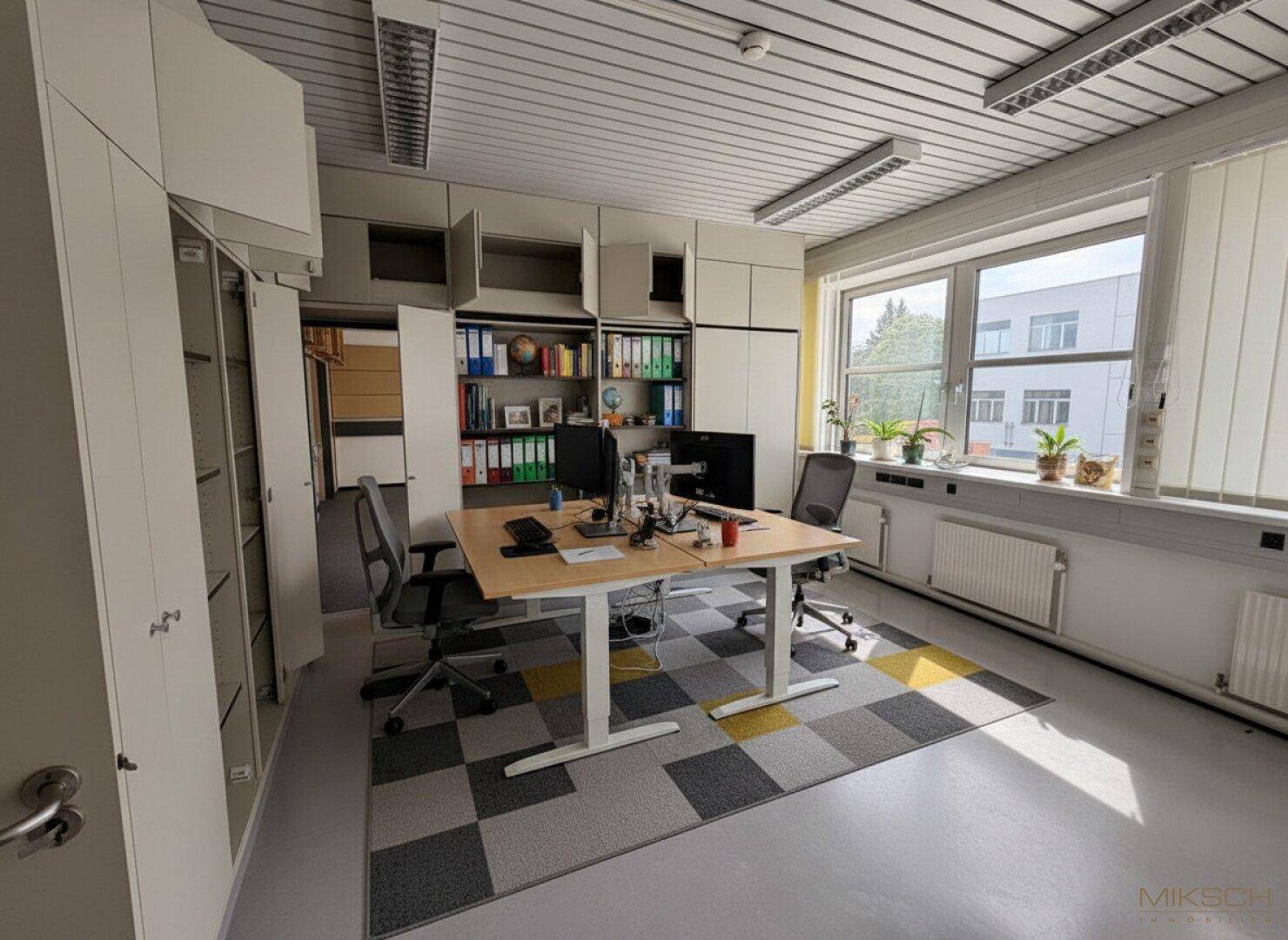 Büro mit Arbeitsplatz, integrierten Schränken und großen Fenstern, die viel Tageslicht hereinlassen.