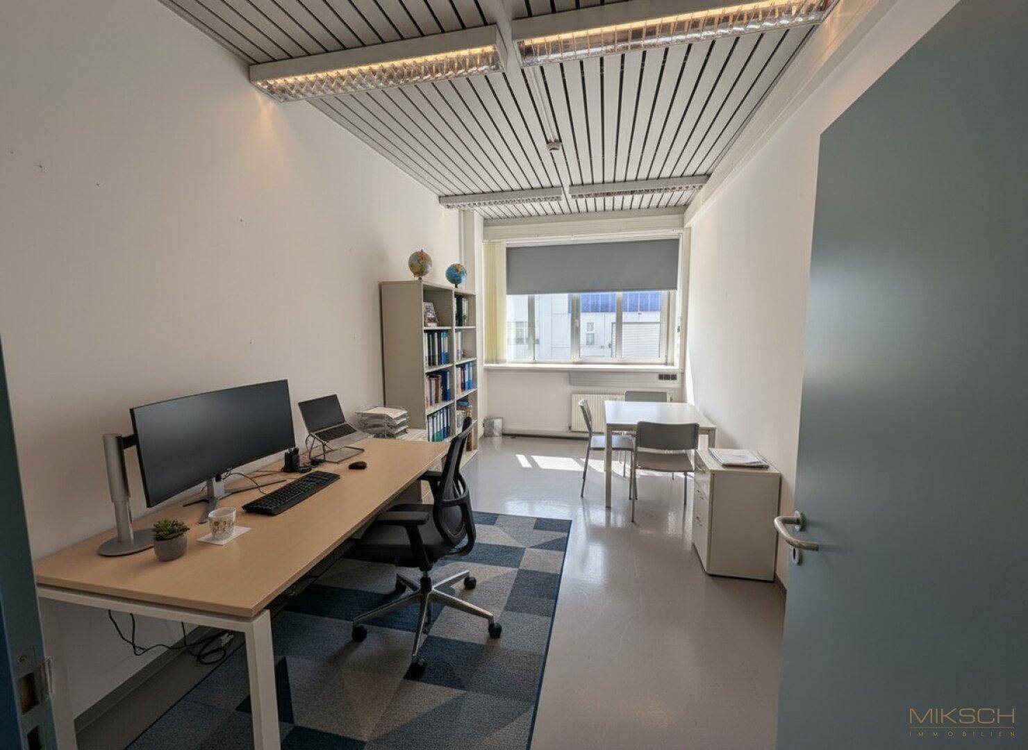 Helles Einzelbüro mit Schreibtisch, Bücherregal und Besprechungstisch, ausgestattet mit einem Rollo am Fenster.