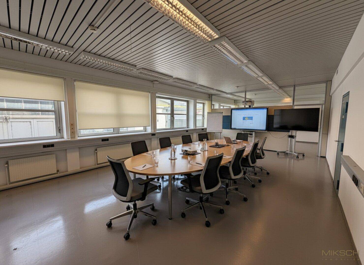 Großer Konferenzraum mit ovalem Tisch, vielen Stühlen und Präsentationsbildschirmen, ideal für Besprechungen.