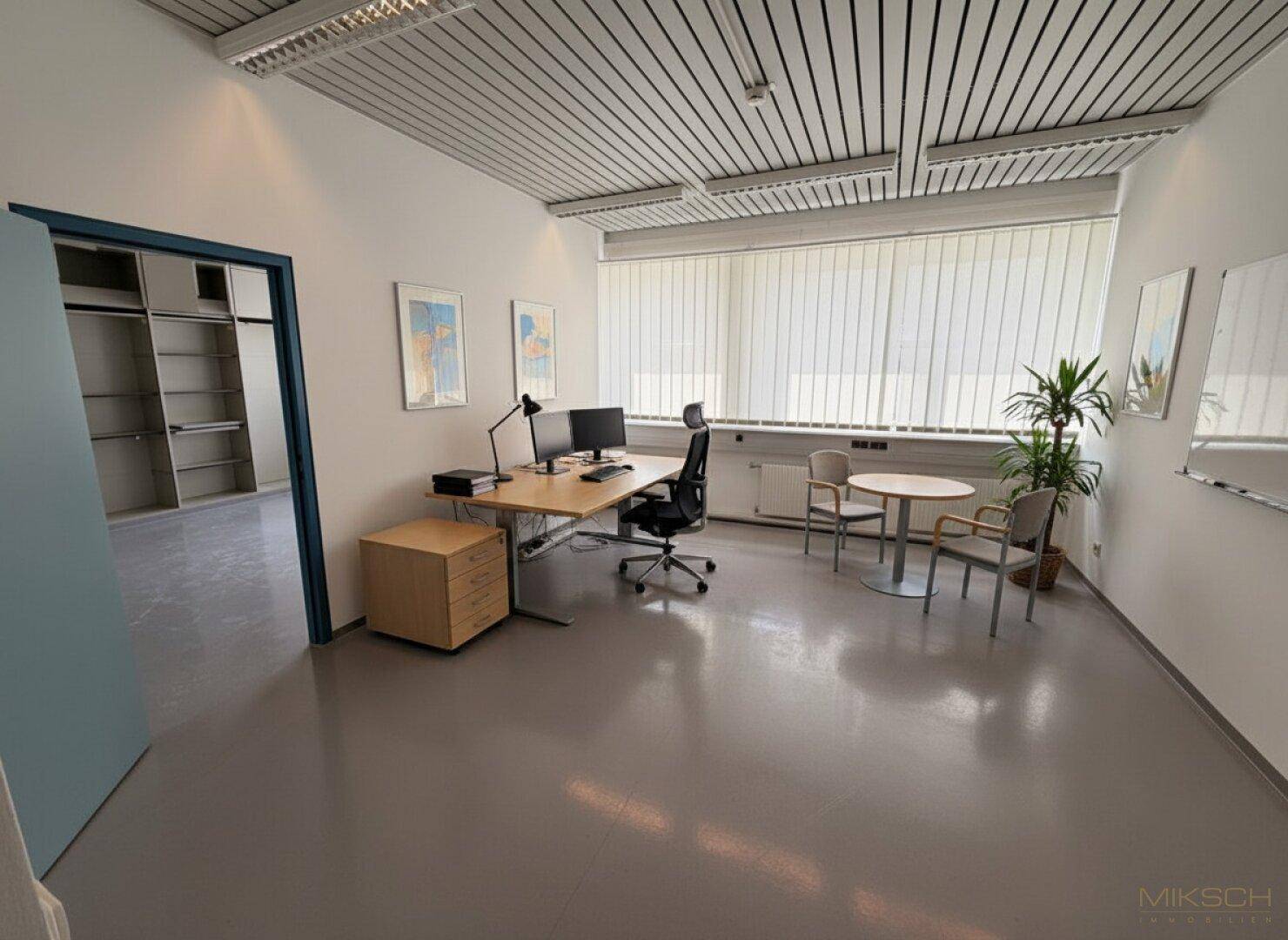 Einzelbüro mit Schreibtisch, Besprechungsecke und Blick nach draußen, angrenzend an einen Raum mit Regalen.