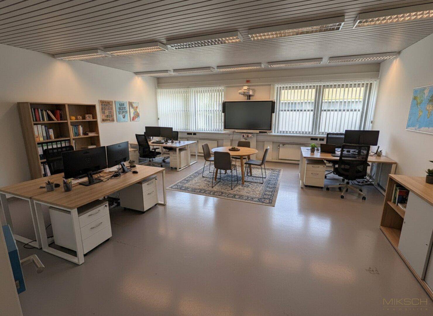 Großes Großraumbüro mit mehreren Arbeitsplätzen, Besprechungsecke und Bücherregalen, hell und funktional eingerichtet.