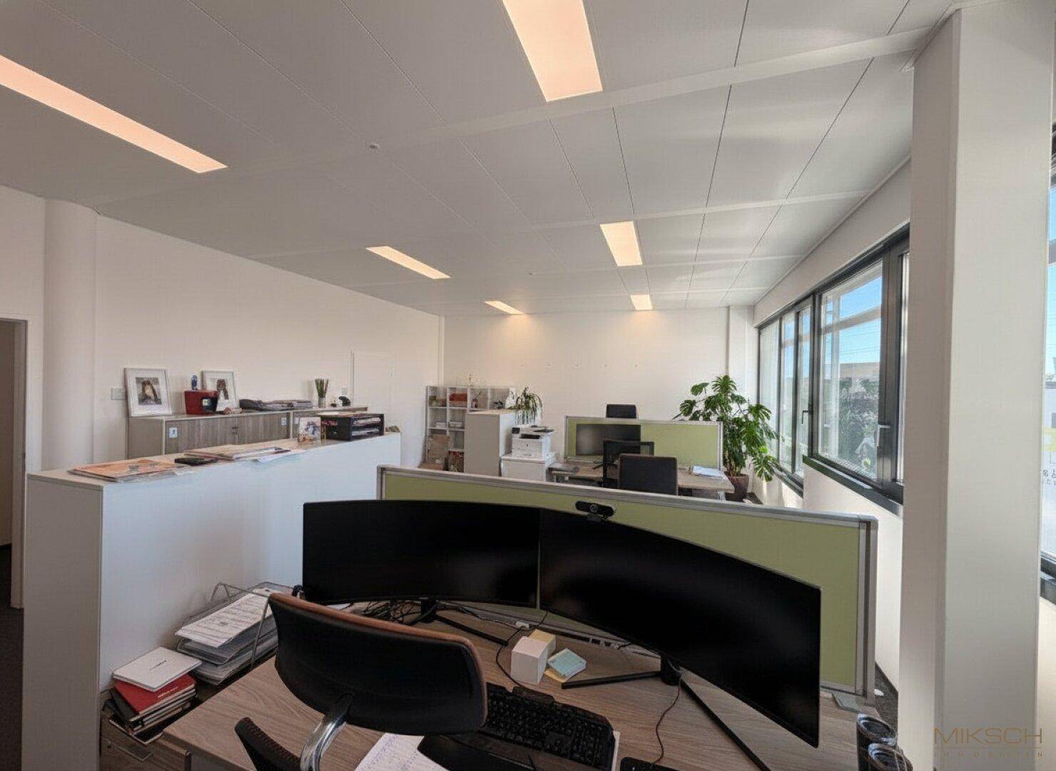 Heller Großraumbüro-Bereich mit mehreren Arbeitsplätzen, großen Fenstern und Teppichboden, gut beleuchtet.