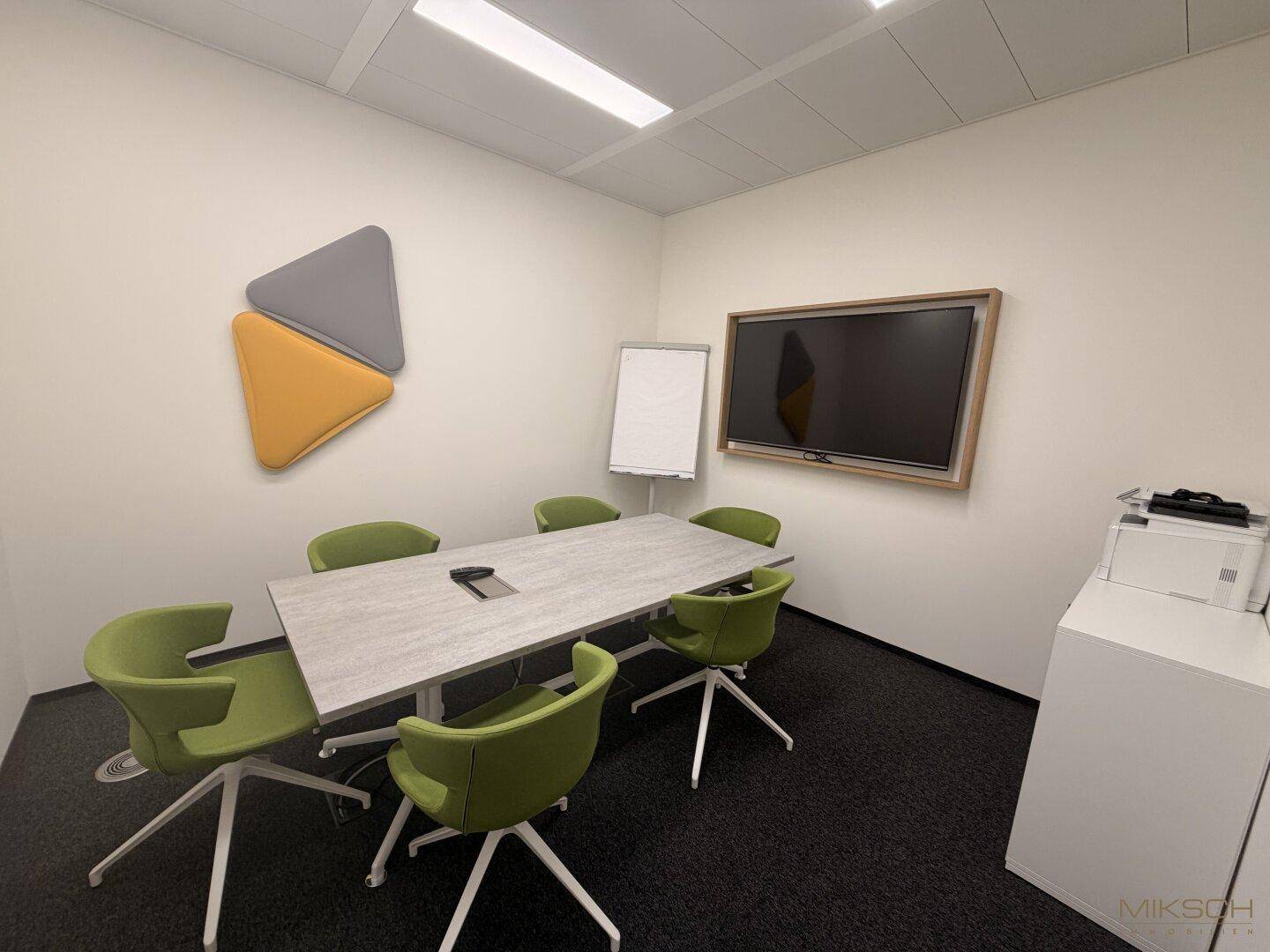Konferenzraum mit modernem Tisch, grünen Stühlen und Wandmonitor für Präsentationen.