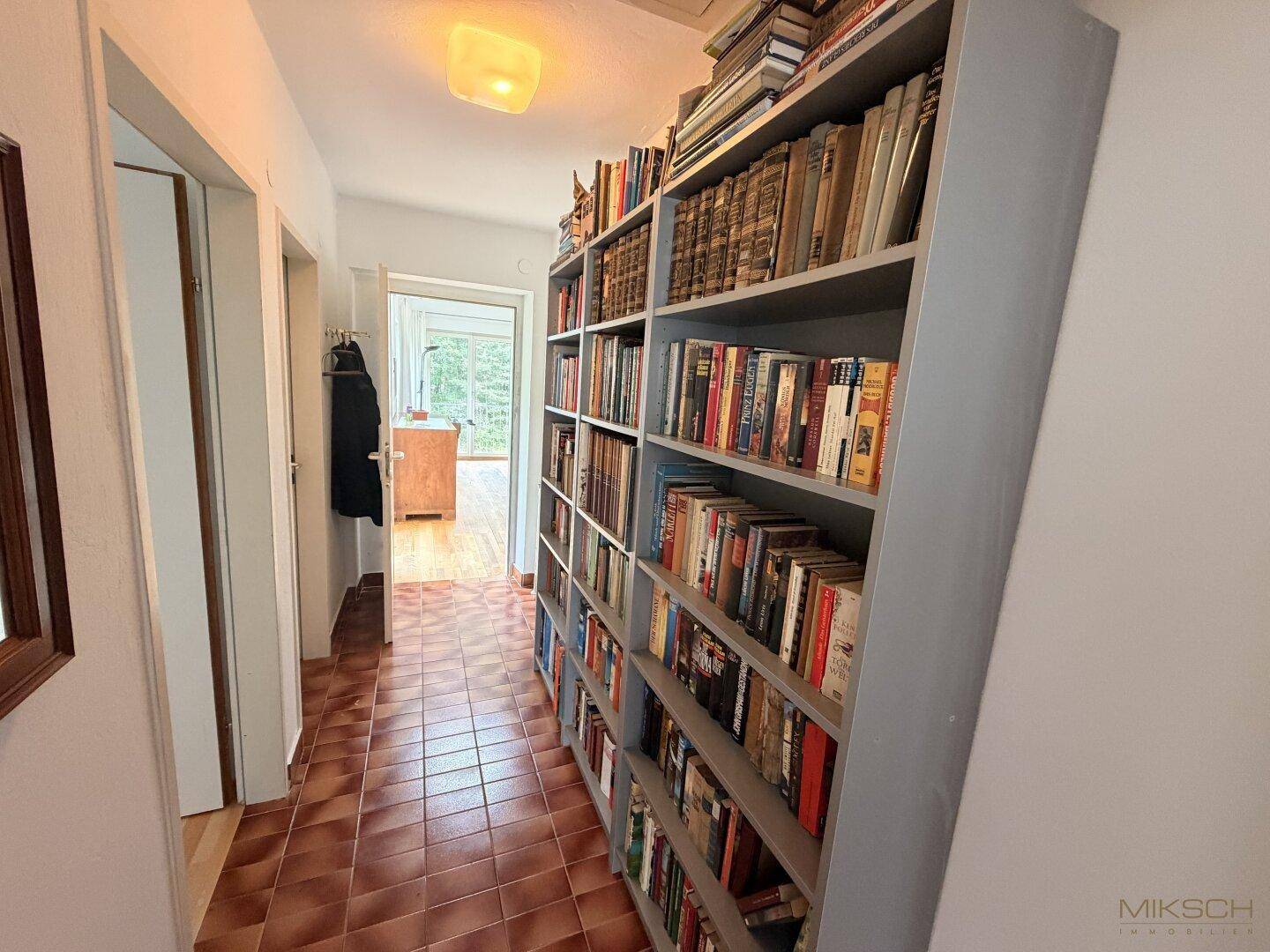 Langer Flur mit einem großen Bücherregal und braunen Fliesen, der zu weiteren Räumen führt.