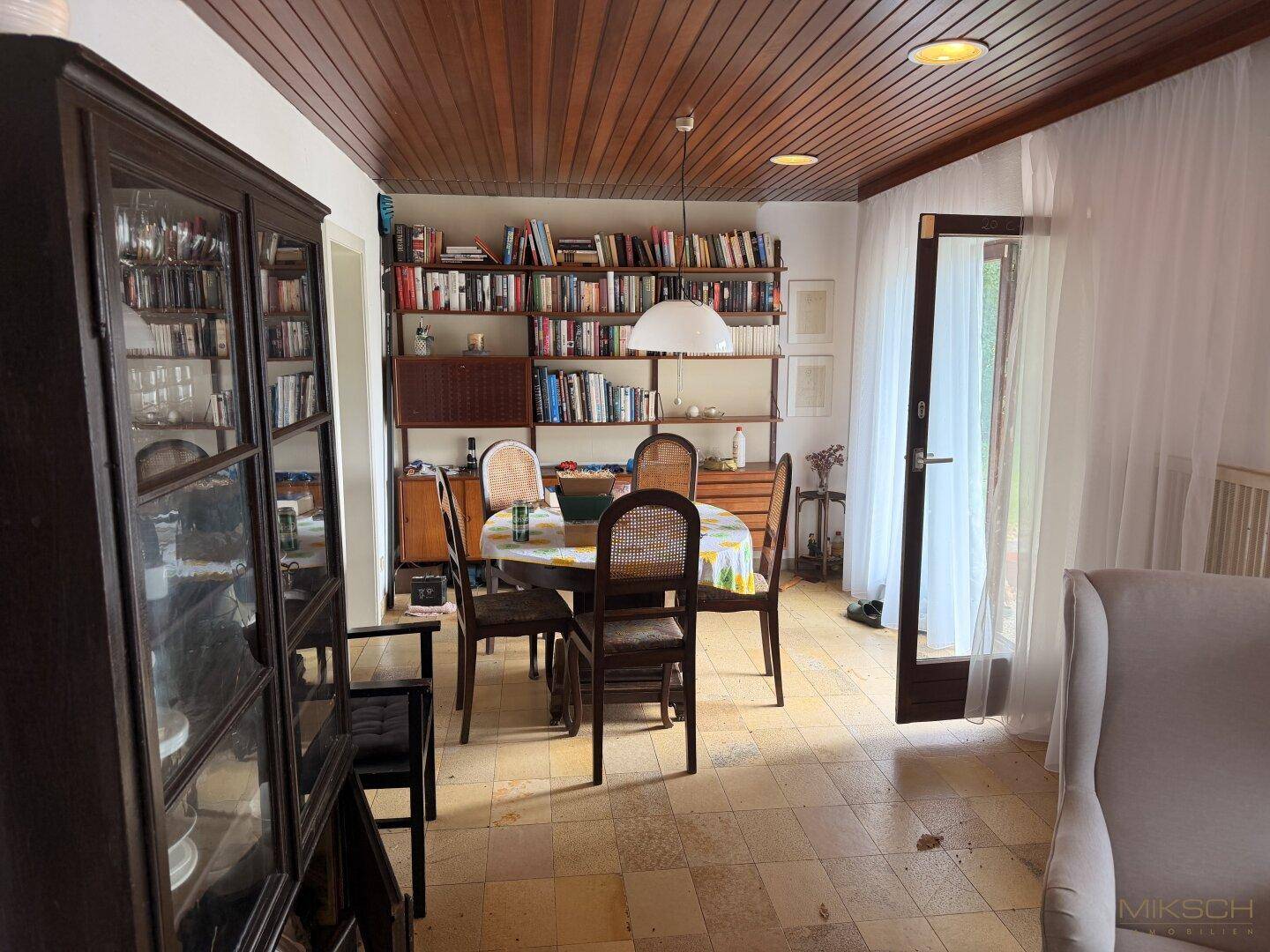 Essbereich mit Holztäfelung an der Decke, runden Esstisch und Bücherregalen, mit Zugang nach draußen.