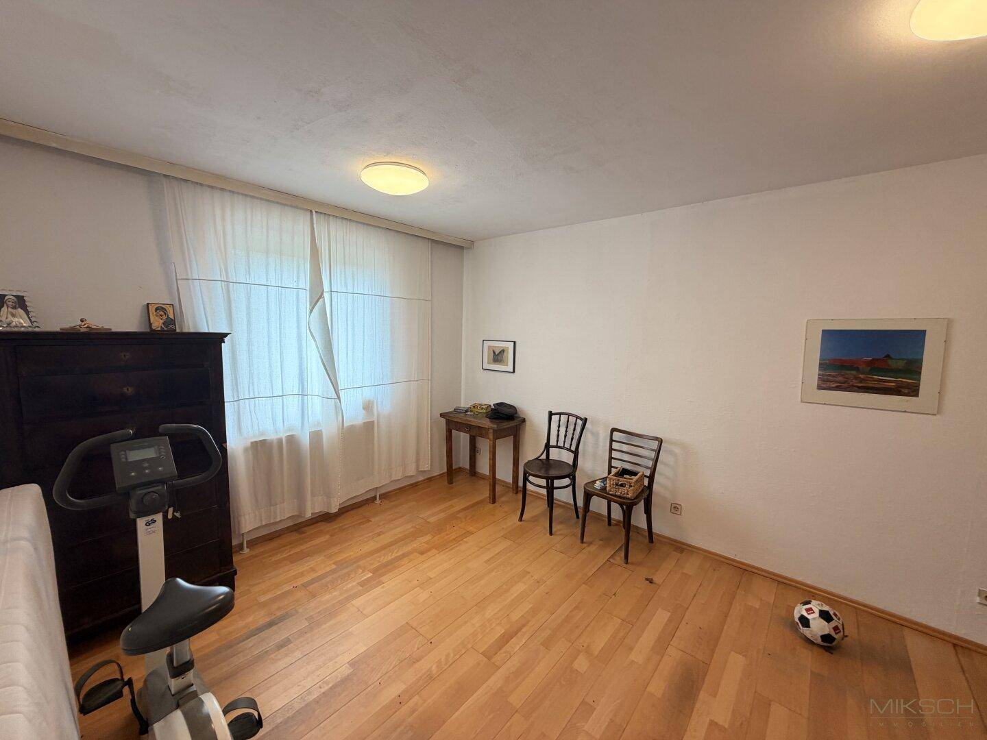 Helles Zimmer mit Holzboden, großem Fenster und einem Heimtrainer, vielseitig nutzbar.