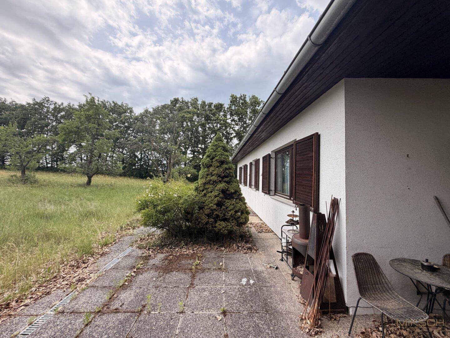 Gepflasterte Terrasse an der Hausseite, umgeben von Rasen und Bäumen, mit Gartenmöbeln.