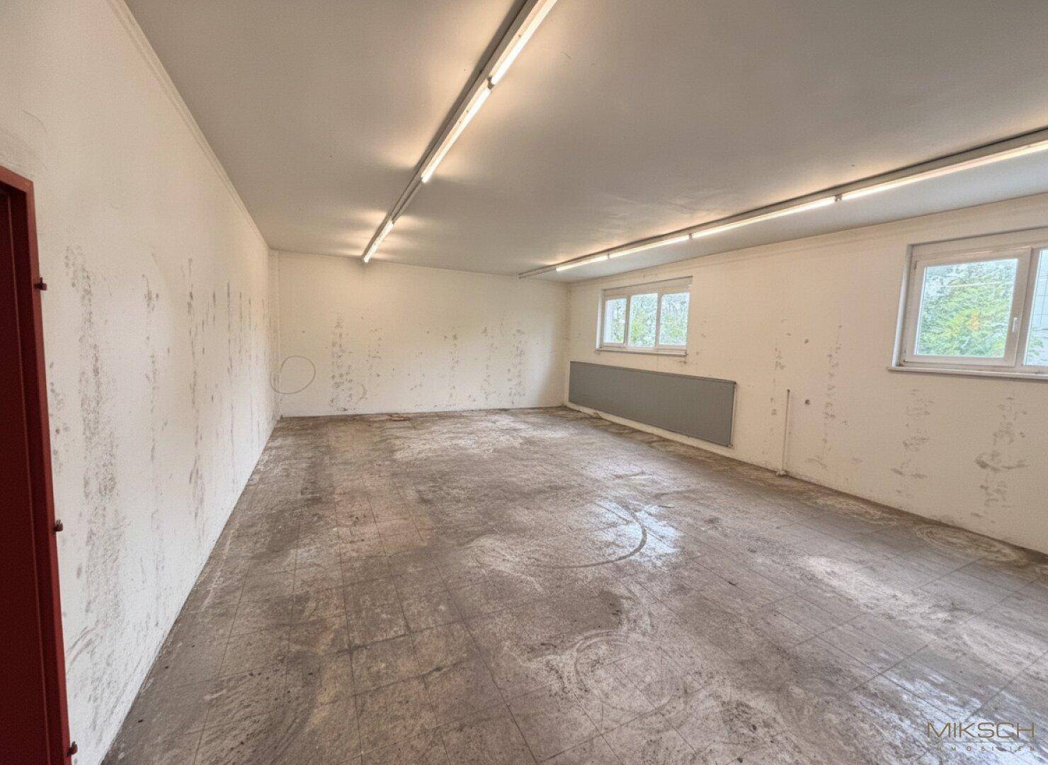 Ein leerer, heller Raum mit Fenstern und einfacher Beleuchtung, nutzbar als Lager oder Büro.