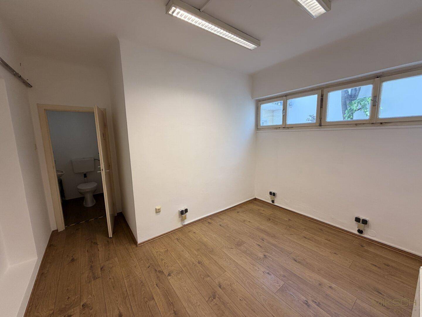 Kleiner Raum mit Laminatboden, weißen Wänden und einem Fenster, der zu einer Toilette führt.