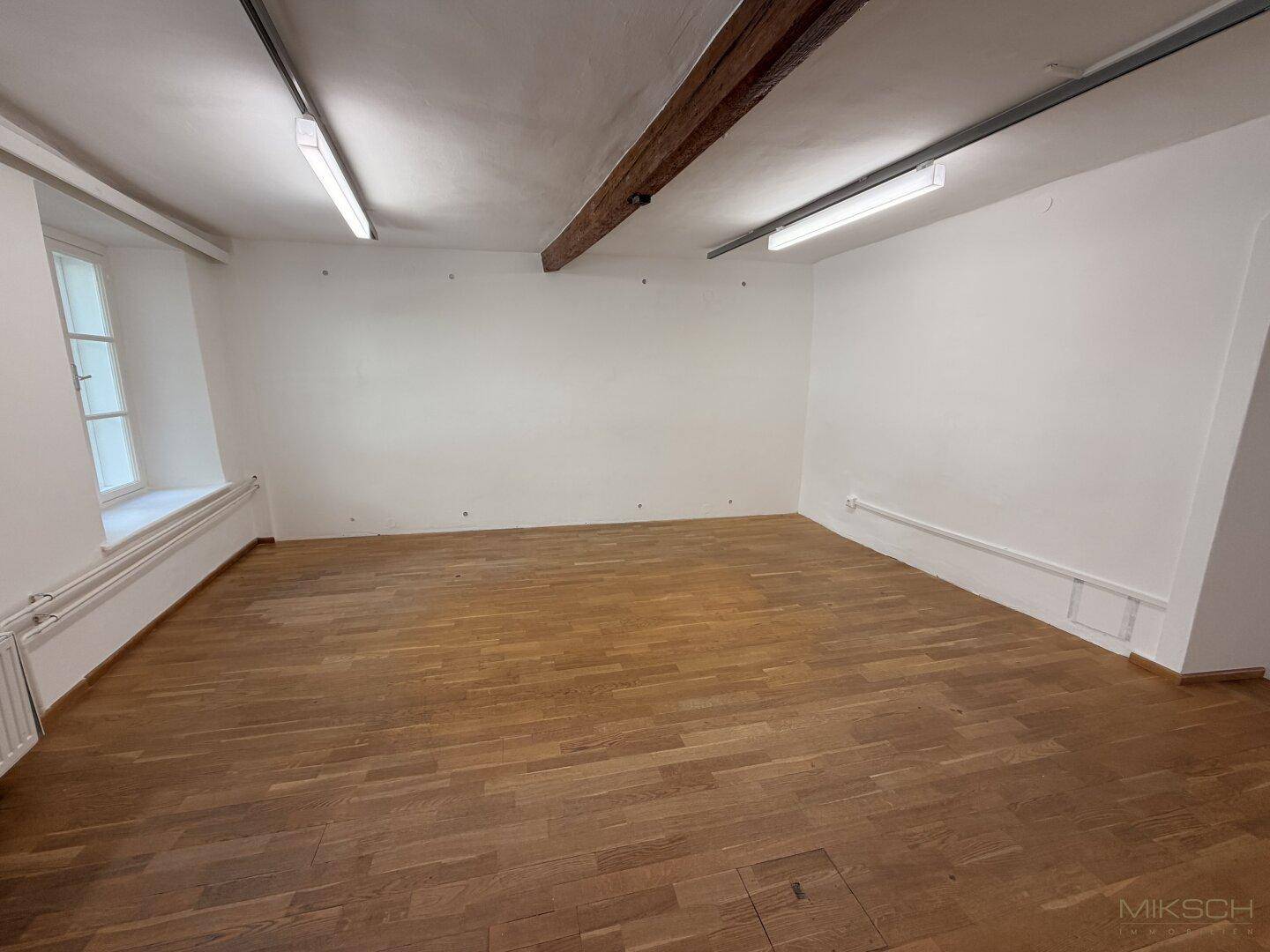Heller Gewerberaum mit Holzboden, weißen Wänden, einem Fenster und sichtbaren Holzbalken an der Decke.